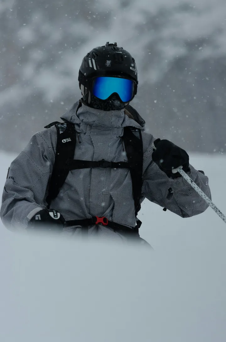 A skier in a gray patterned jacket, black helmet, and blue goggles in falling snow.