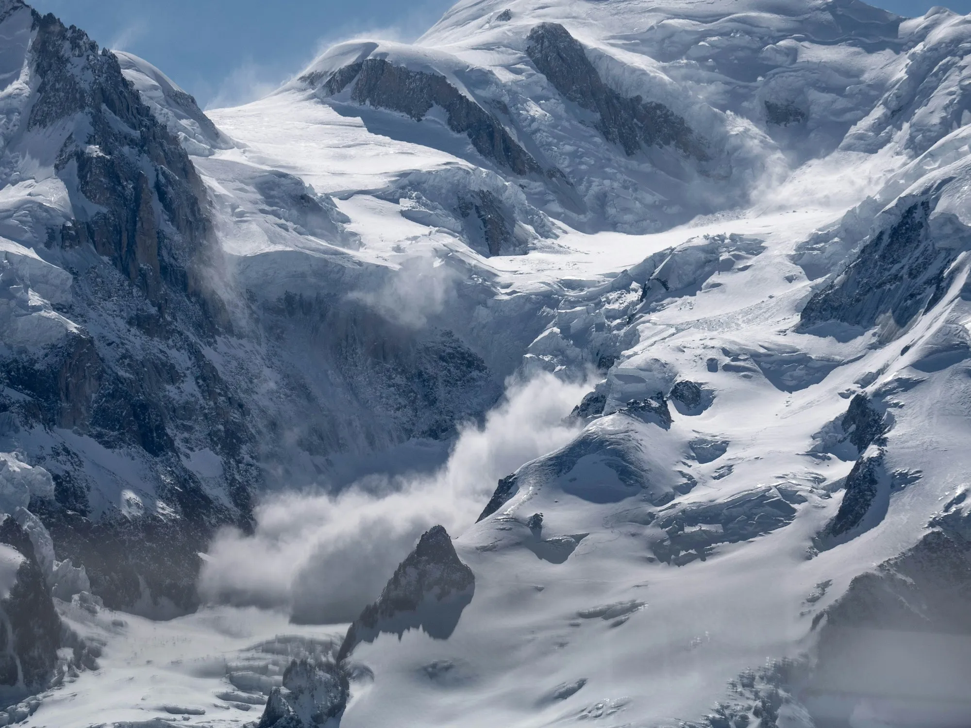 A powder-snow avalanche cascading down a steep alpine ridge under blue sky, glaciated peaks above.