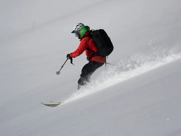A skier in a red jacket and black backpack carving through soft snow on a steep slope, spray kicking up behind.