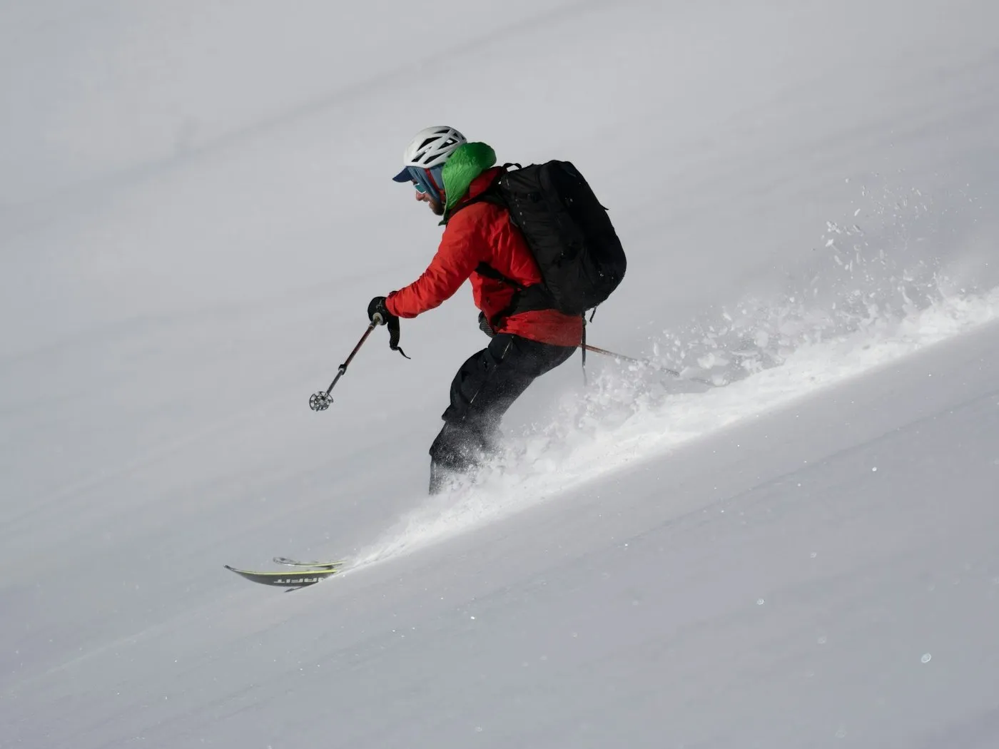 A skier in a red jacket and black backcountry backpack carving through soft snow on a steep open slope, spray kicking up behind.