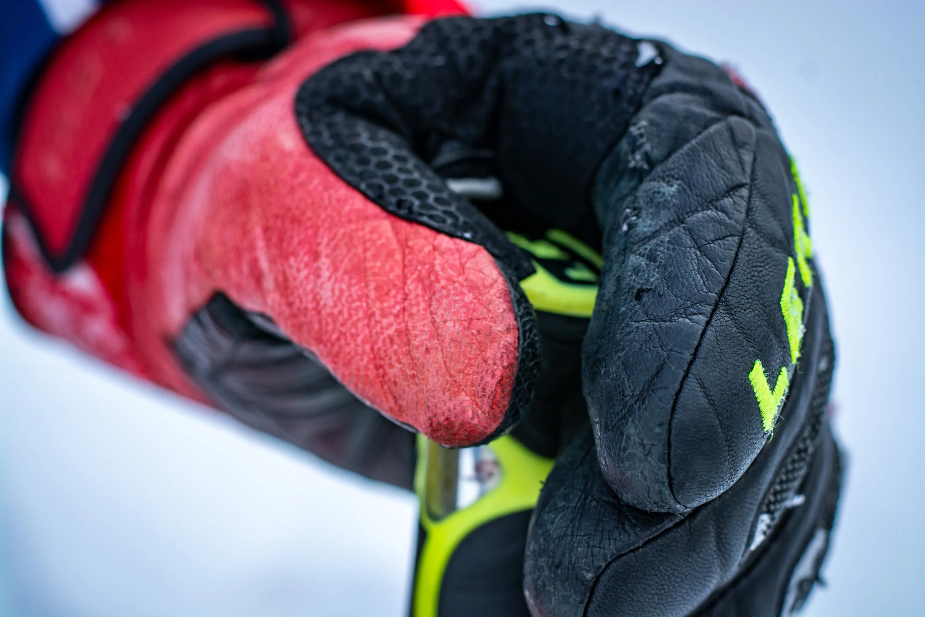 Close-up of two worn leather ski gloves, one red and one black with neon-yellow accents, gripping ski poles in snow.