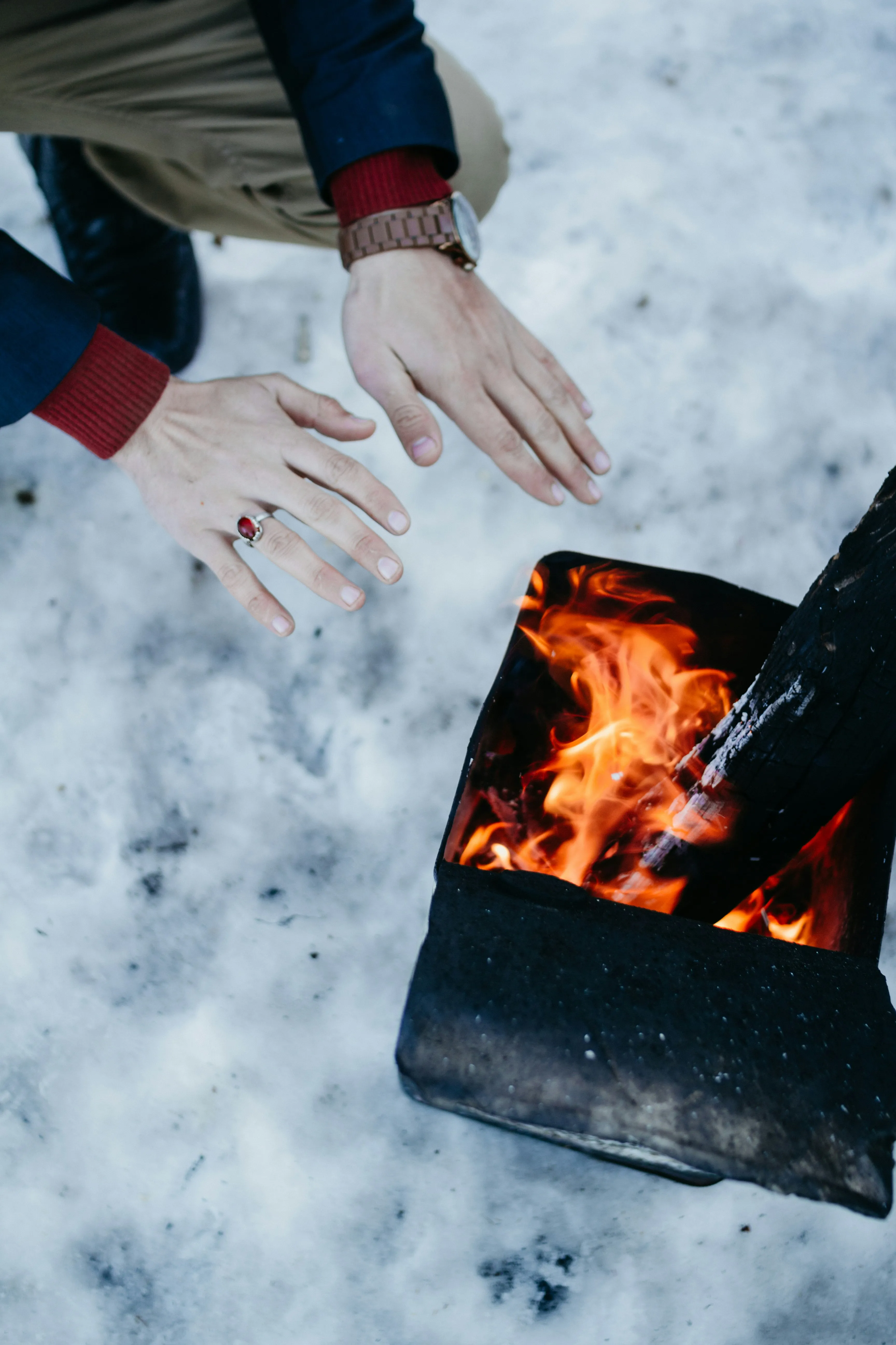 A person warming bare hands over a small open fire in a black metal fire pit set on snow.