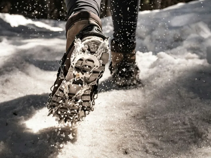 Close-up of a hiking boot fitted with a strap-on ice-traction cleat, chains and spikes across the sole, kicking up snow.