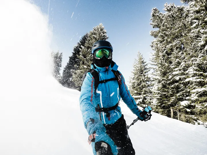 A skier in a blue jacket, black helmet, and green goggles skiing through powder beside snow-covered trees.
