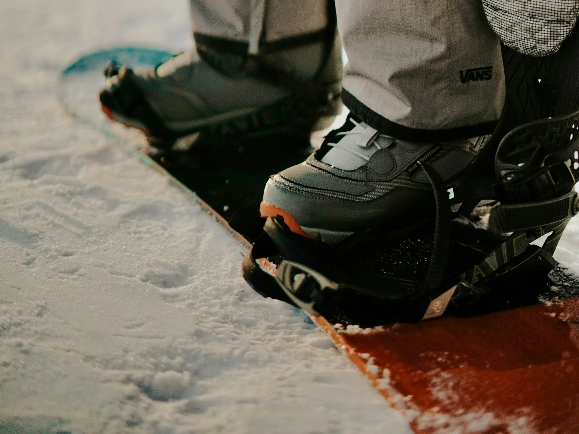 Close-up of a snowboard boot strapped into a binding on a board, laces and binding straps visible, a second boot behind, snow all around in low golden light.