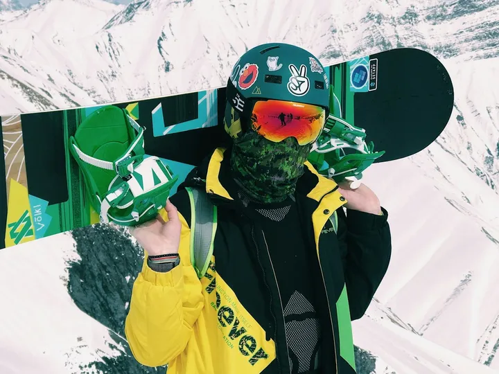 A snowboarder in a sticker-covered helmet and orange-mirrored goggles carrying a green board, snowy peaks behind.