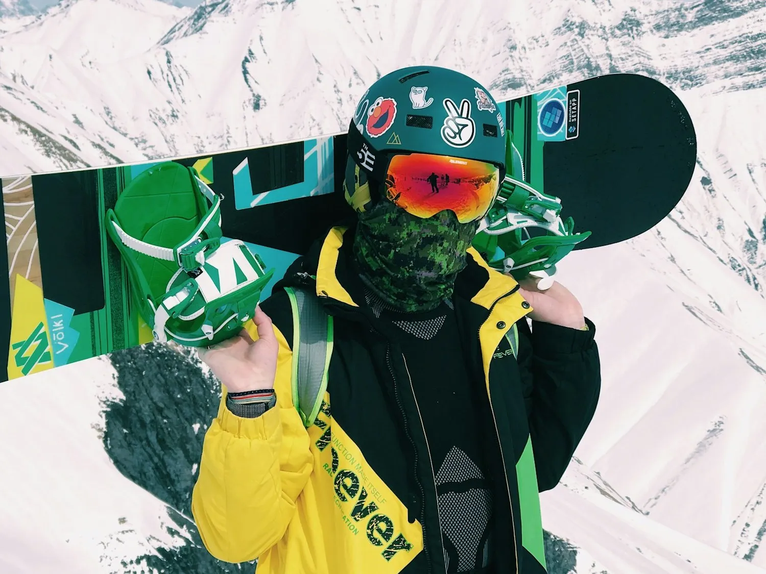A snowboarder in a sticker-covered helmet and orange-mirrored goggles with a camo neck gaiter, carrying a green Völkl board across the shoulders, snowy peaks behind.