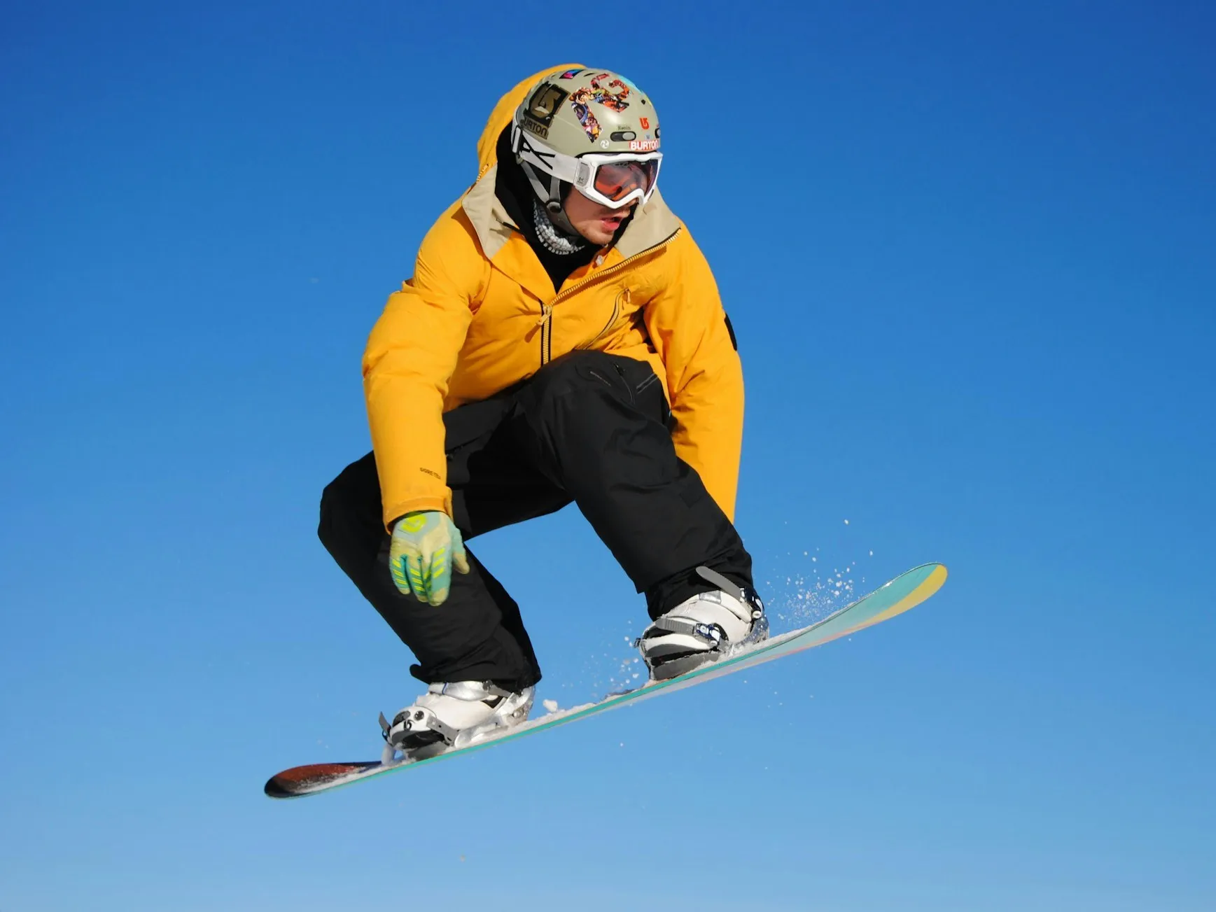 A snowboarder in a bright yellow jacket and black pants grabbing the board mid-jump against a clear blue sky.