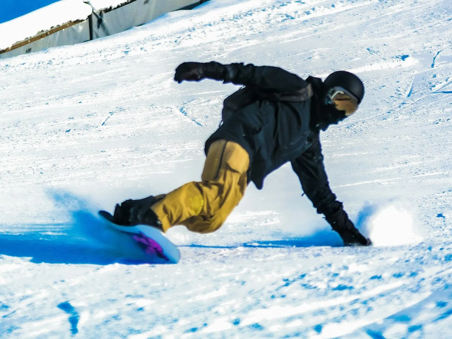 A snowboarder in gold-yellow pants and a black jacket carving hard and low with a hand dragging in the snow, the board spraying, on a sunlit slope.