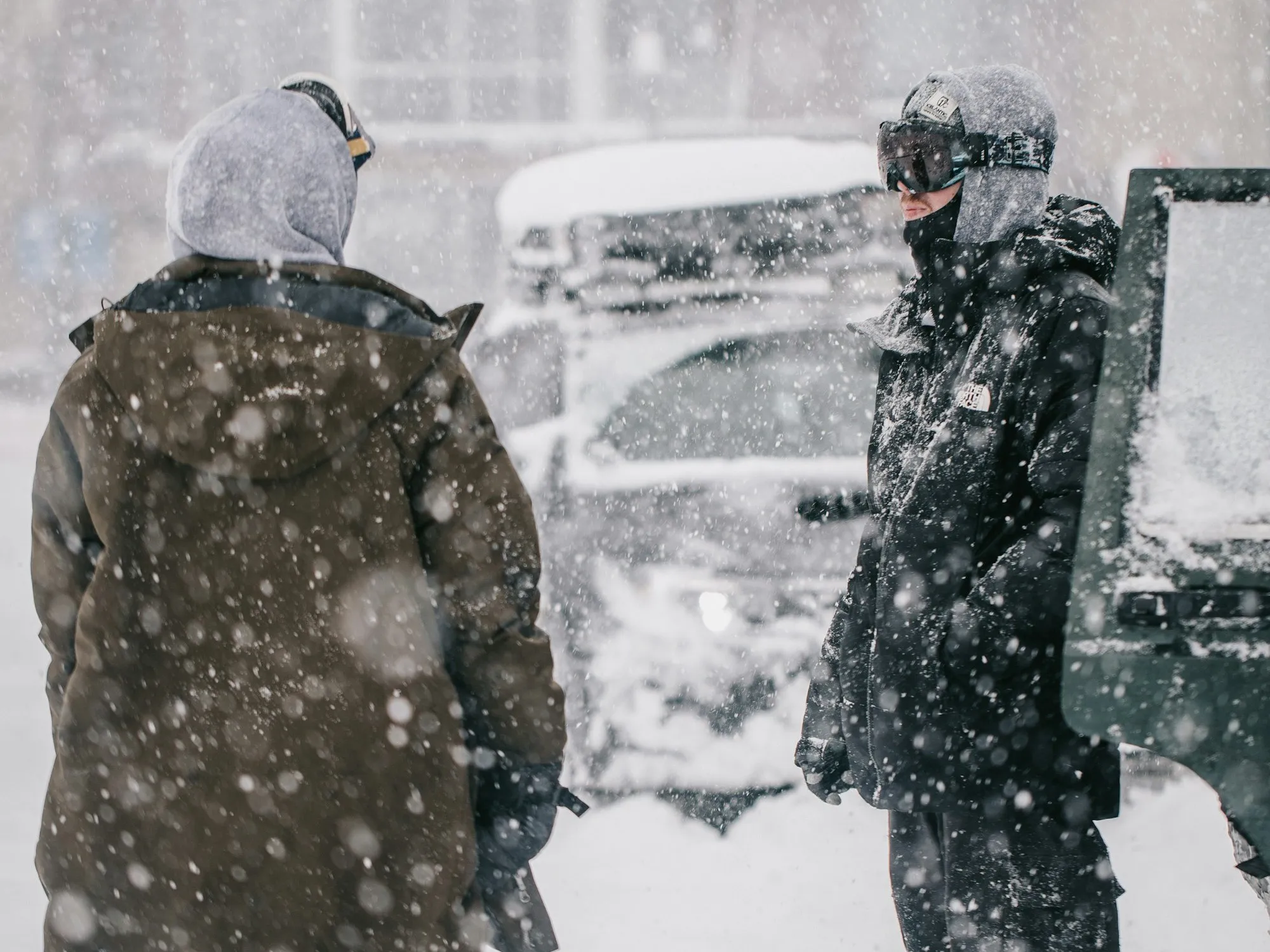 Two people in heavy hooded parkas standing in driving snow beside a snow-buried car during a blizzard.