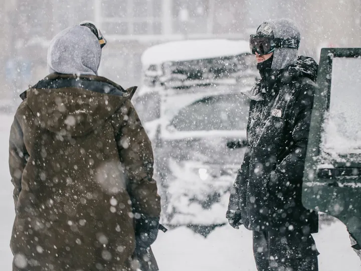 Two people in heavy hooded parkas standing in driving snow beside a snow-buried car during a blizzard.