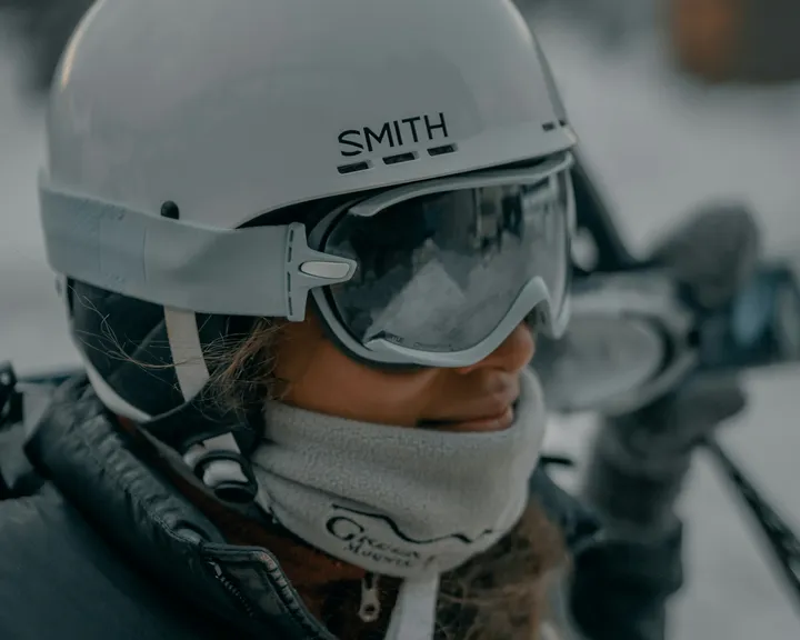 A skier in a white Smith helmet and mirrored Smith goggles with a fleece neck gaiter, ski poles behind.