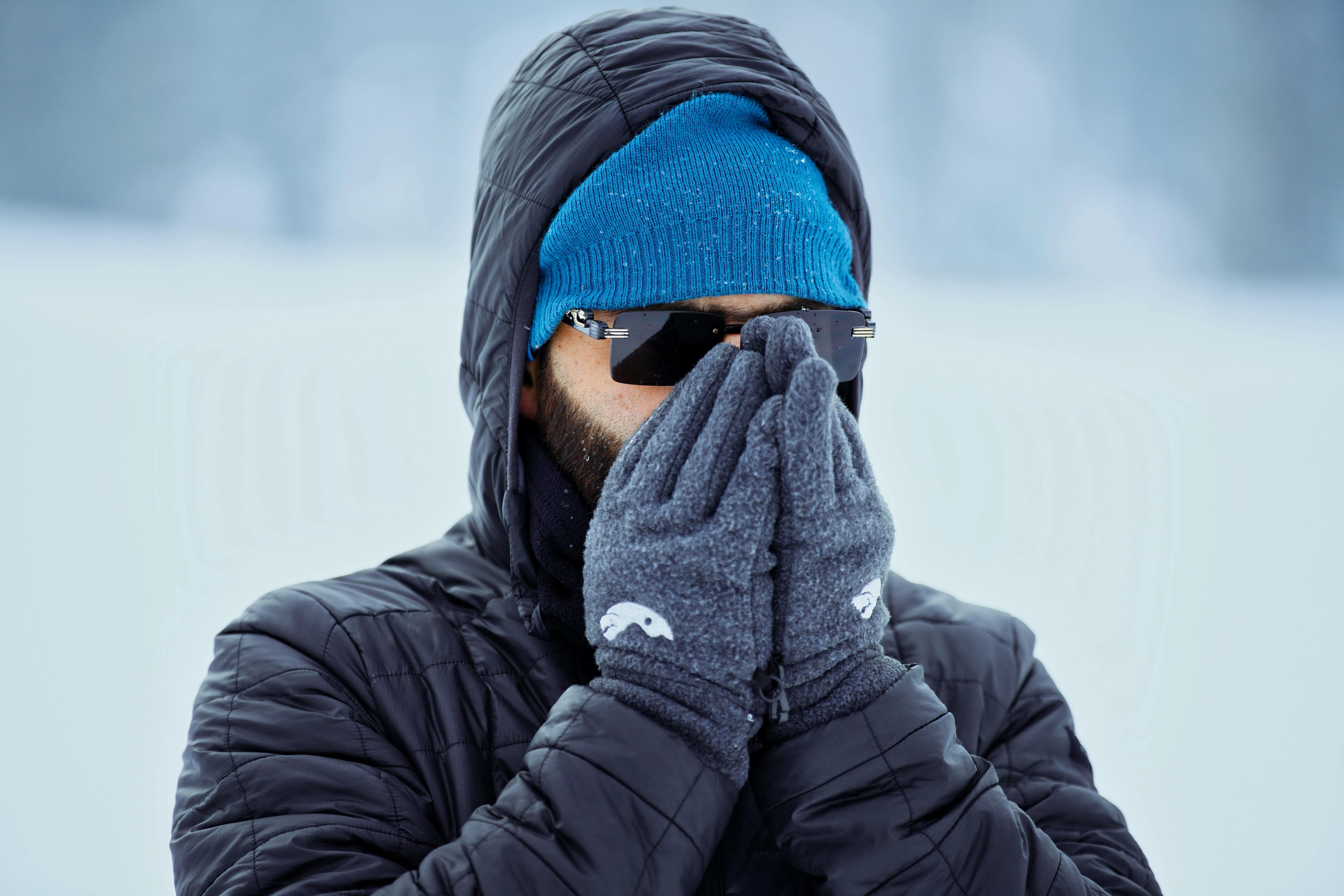 A man in a black hooded puffer, blue knit beanie, sunglasses, and gray fleece gloves held to his face in the snow.