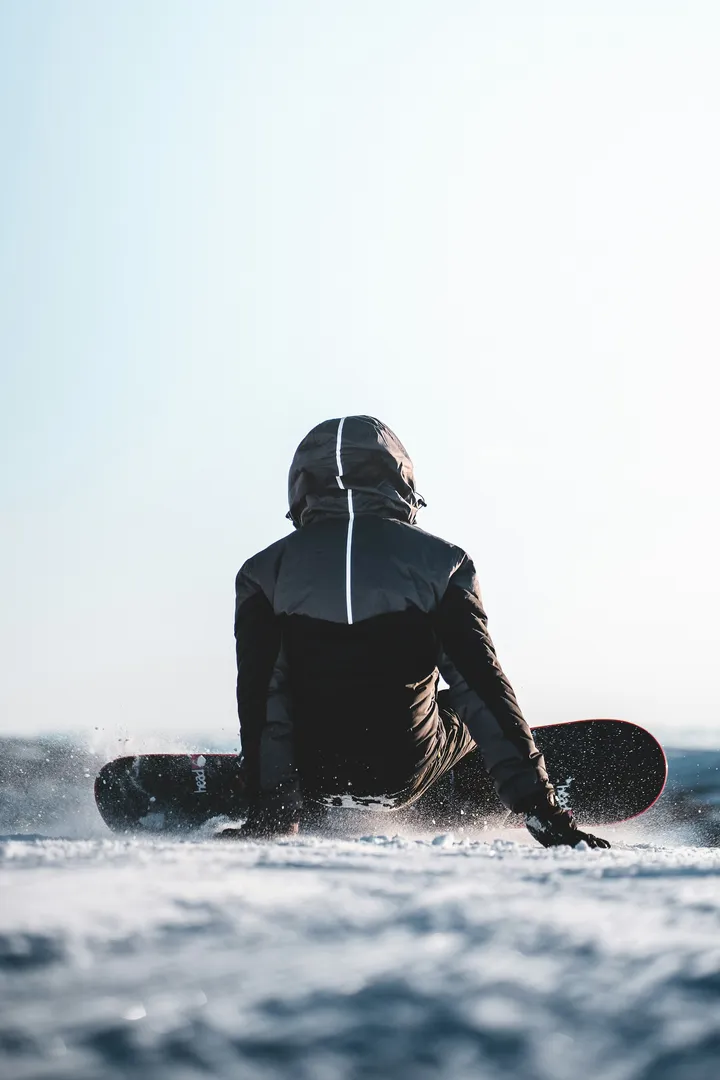 A snowboarder sitting back into a turn on a bright day, snow spraying off the edge, seen from behind.