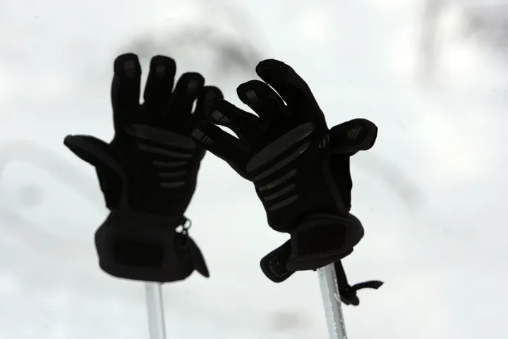 A pair of black insulated ski gloves resting fingers-up on ski poles against a snowy backdrop.