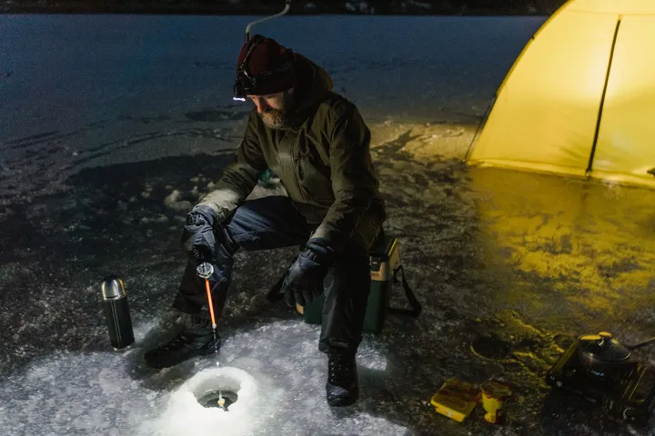 A man in a headlamp kneeling on a frozen lake at night, jigging a short rod over an ice hole, a lit tent beside him.