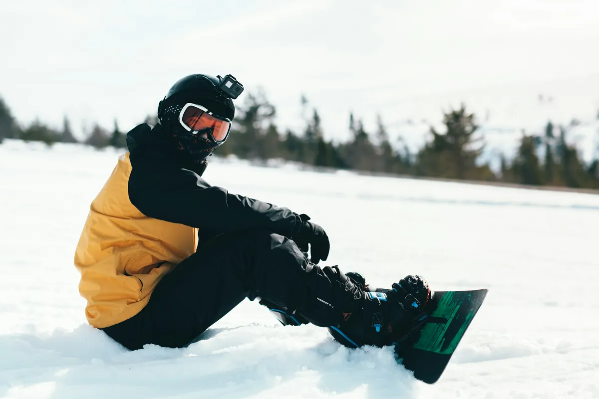 A snowboarder in a yellow and black jacket and black pants sitting in the snow with boots strapped into a snowboard.