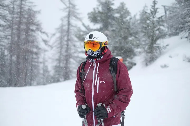 A skier in a burgundy jacket, white helmet, and orange goggles standing in falling snow.