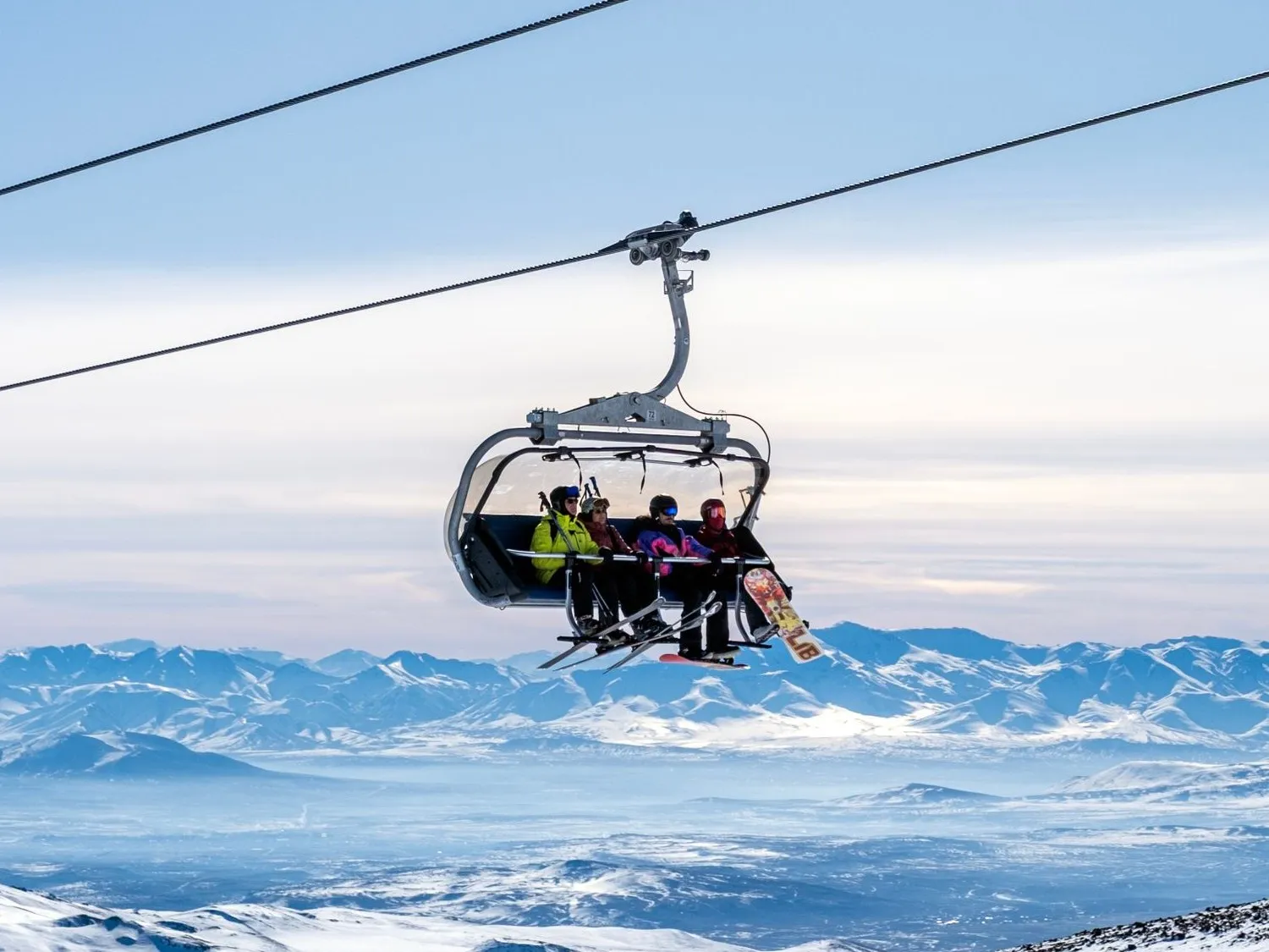 A four-seat detachable chairlift carrying skiers and a snowboarder over a vast snowy mountain range under a clear sky.