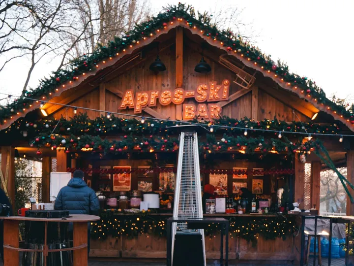 An outdoor apres-ski bar at dusk, a lit 'Apres-Ski Bar' sign over a wooden chalet counter with fairy lights.
