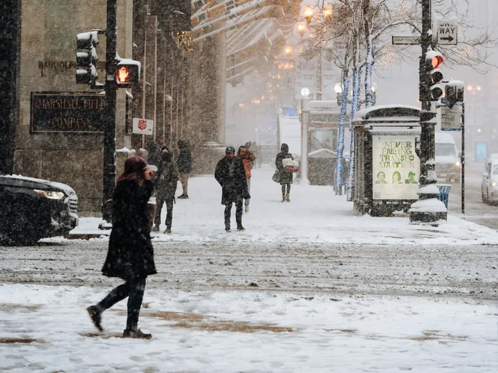 Pedestrians in heavy coats crossing a snowy downtown Chicago street, the historic Marshall Field building behind.