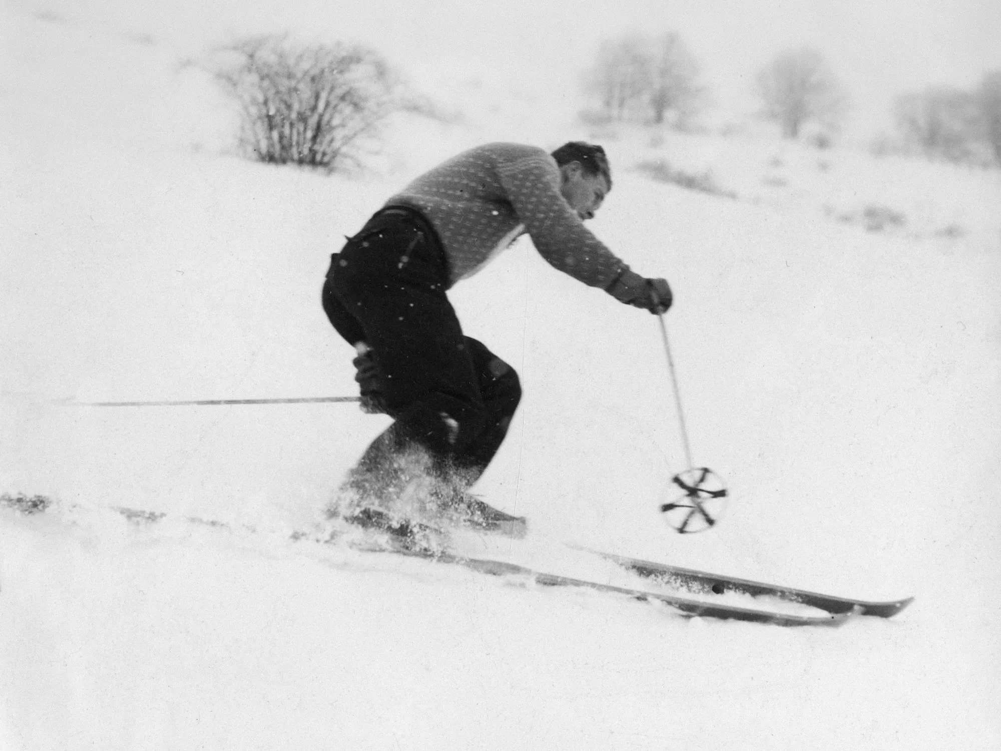 A vintage black-and-white photo of a skier in a patterned knit sweater carving downhill on long wooden skis with a large basket pole, snow spraying underfoot.
